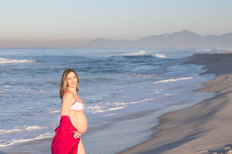 Gestante de vestido rosa caminhando na beira da praia no início da manhã