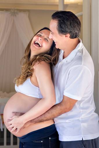 casal de gestantes abraçados sorrindo em frente ao quarto do bebê.