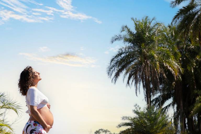 Gestante sorrindo em ensaio com coqueiros ao fundo na praia.