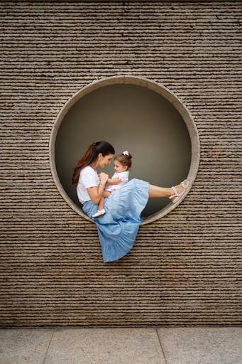 Mãe sentada dentro de uma moldura circular em uma parede com sua filha bebê no colo; Mae e filha conectadas e interagindo, uma de frente para a outra; roupas claras, tons neutros