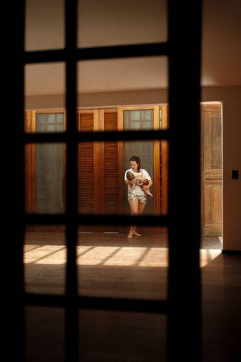 Mãe segurando bebê no colo em uma sala iluminada pela luz natural do sol; silhueta da janela forma uma moldura sobre a mãe e o bebê; tons quentes 