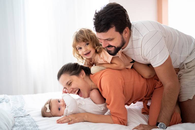 Família deita em montinho uns sobre os outros na cama; bebê por baixo, mãe e filha no meio e pai abraçado por cima; foto alegre e espontânea; todos sorrindo; meninas