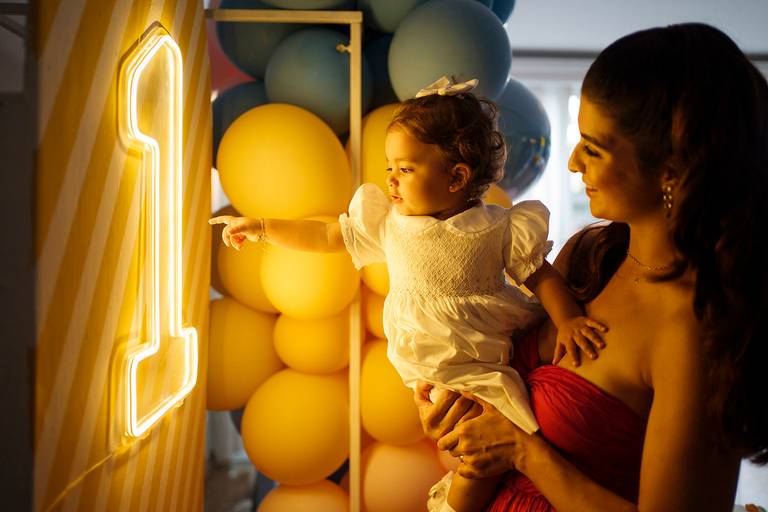 Mãe segurando bebê em festa infantil no colo; primeiro aniversário do bebê; aniversário de 1 ano; um ano; bebê apontando para placa luz de Led iluminada; tons quentes