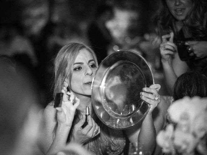 Foto no estilo documental feita em uma festa de casamento , na cidade de São Paulo capital, onde uma convidada usa um aparador de prato reflexivo, de metal para retocar o batom 
