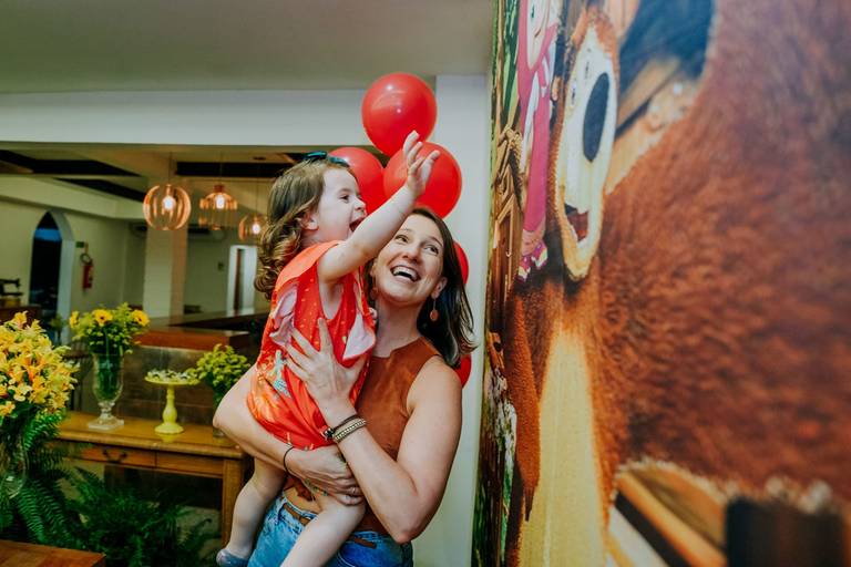 menina de 3 anos no colo da tia se diverte batendo no painel da masha e o urso que decora sua mesa de bolo