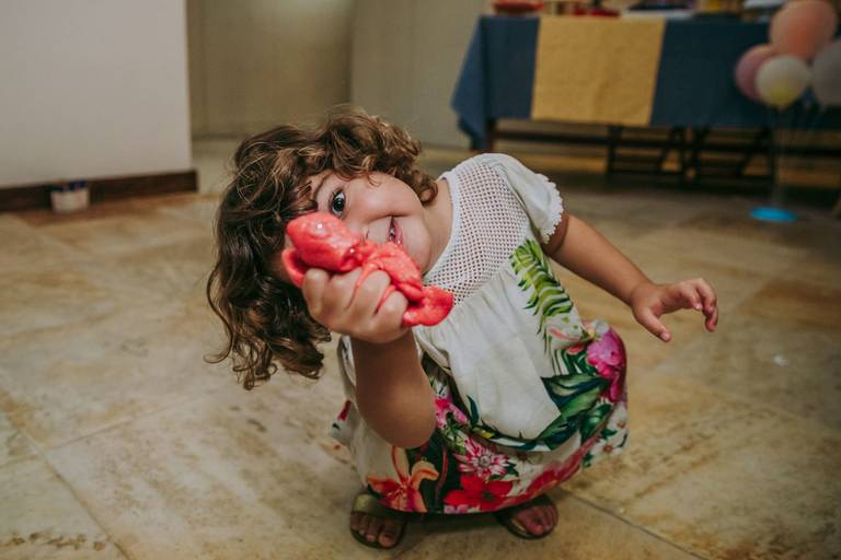 menina de tres anos com slime na mão e cobrindo a metade do rosto sorri carinhosamente  para a camera