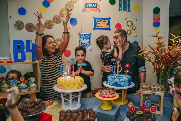 foto da hora do parabéns, mae  animada e com os braçoslevantados, filho menor batendo palmas, filho maior segura o rosto do pai que faz bico pra ele