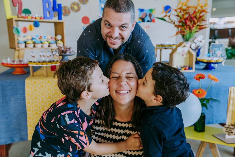 dois filhos beijam cada um um lado do rosto de sua mae que esta de olhos fechado enquanto seu pai sorri