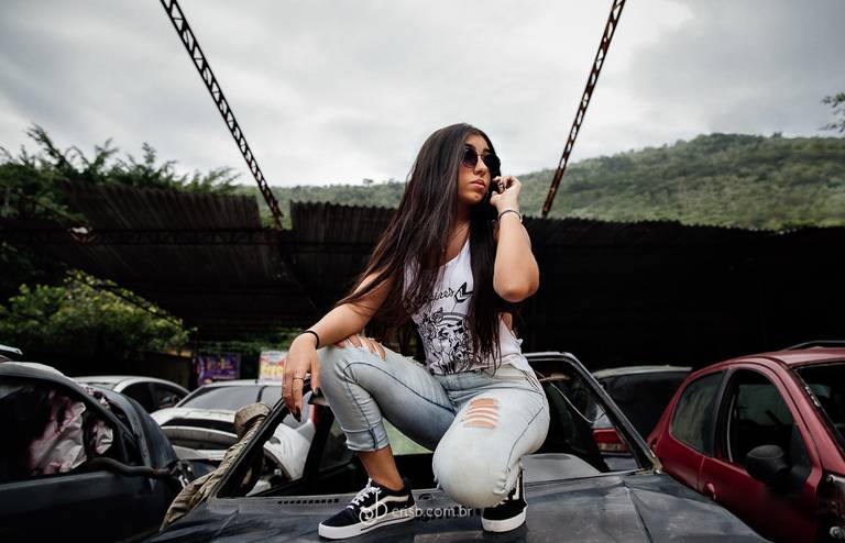 book de debutante fotografada dentro de ferro velho na estrada do catonho no rj debutante está agachada sobre um carro