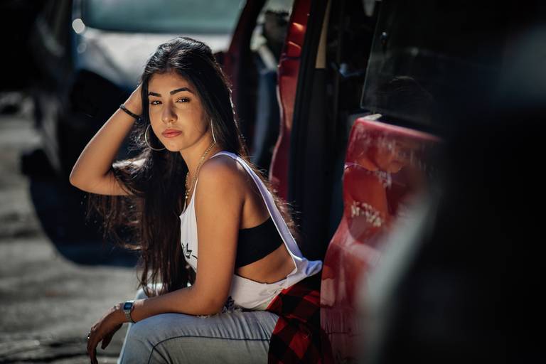 foto lateral de debutante sentada em carro vermelho no ferro velho com a mão direita ajeitando o cabelo
