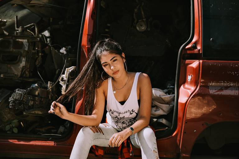 menina sentada na lateral do carro vermelho sem portas ela segura uma mecha do cabelo enquanto olha séria para a camera