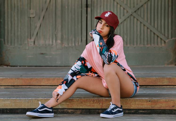 foto de debutante sentada no chão da praça mauá ela está de bone, casaco salmão, short e tenis