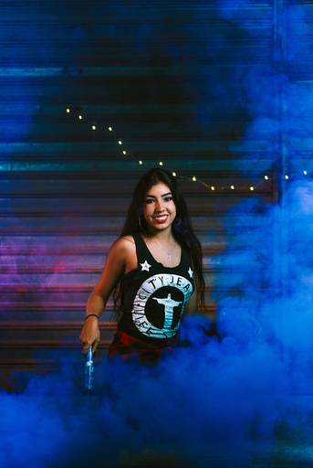 menina com fumaça azul acesa sorrindo para a camera e agachada no chão, atrás dela uma porta de loja sendo iluminada pelas luzes rosa e azul