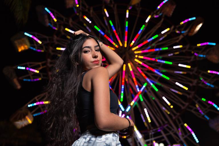 foto noturna tirada de baixo pra cima com uso de softbox em que menina em primeiro plano colocando a mão esquerda ao cabelo e ao fundo está uma roda gigante acesa