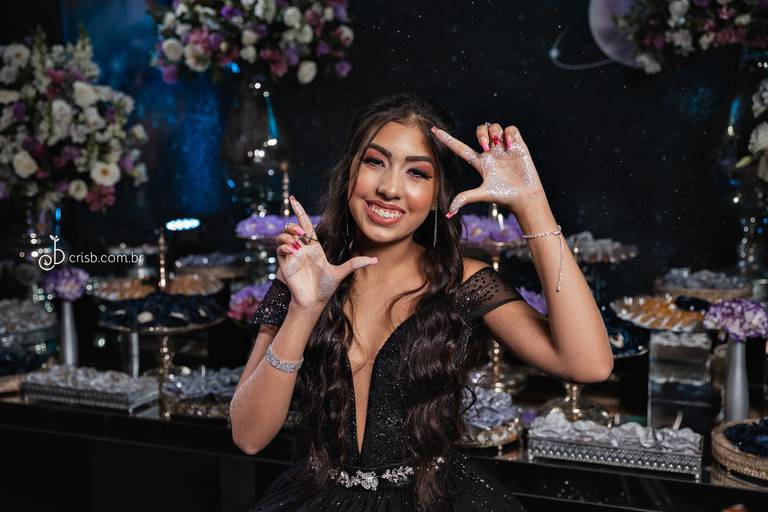 debutante fazendo modulra no rosto com ads mãos sujas de glitter ela de vestido preto em frente a mesa do bolo na casa de festas svm eventos em piedade