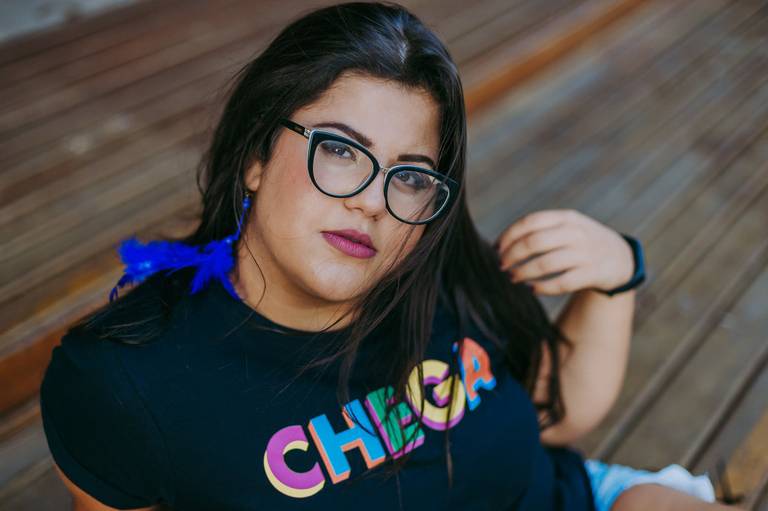 foto de menina de oculo com uma camisa escrito chega ela segura uns fios de cabelo enquanto olha pra camera