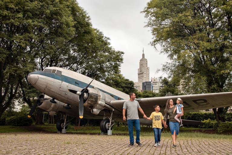 Família caminhando na área externa  do Museu Catavento. Eles estão  estão  de mão dadas, o pai, o filho e a mãe está com filha menor no colo. Estão próximos ao famoso avião. Bem ao fundo podemos ver a torre do Farol Santander.