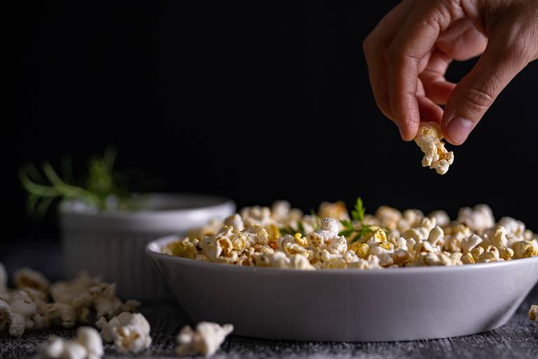 Pipoca em um prato branco com um ramequim ao fundo com temperos. Uma mão feminina está pegando uma pipoca. O prato contém ramos de cheiro verde decorando. A base é de madeira rústica de demolição e o fundo preto.