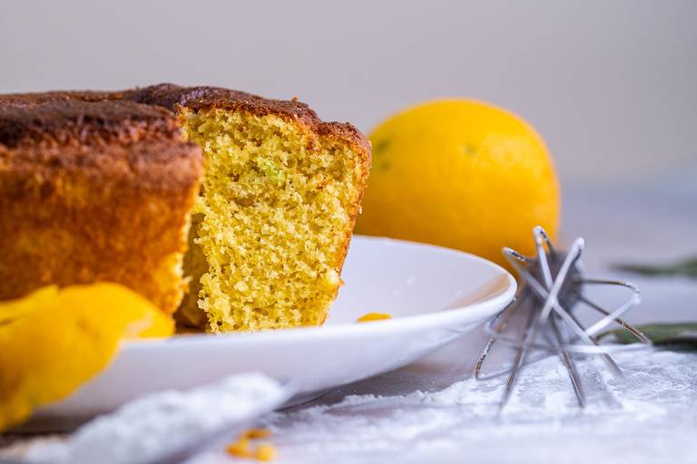 Um bolo sabor laranja inteiro com duas laranjas ao fundo, algumas folhas verdes, uma pedaço de casca de laranja à esquerda, uma colher com farinha e um fuê à direira. A base é uma pedra de mármore nas cores branca e rosa.