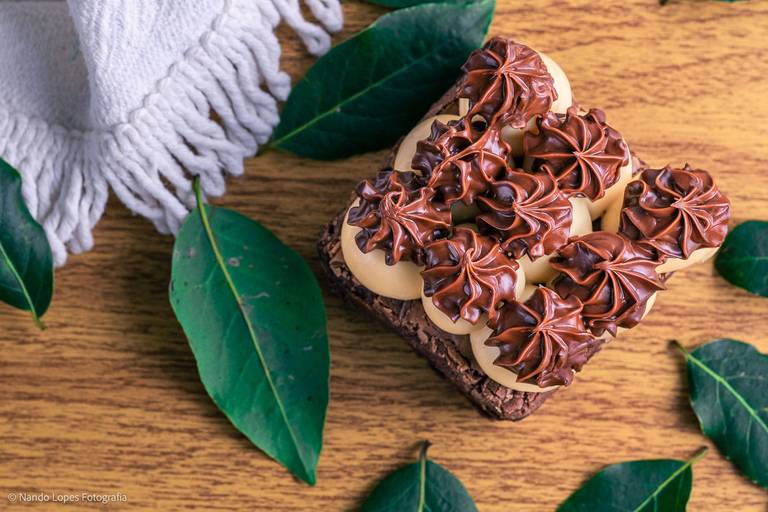 A sobremesa doce é um pedaço de bolo tipo brownie recheado com brigadeiro branco e confeitado de brigadeiro tradicional. Ao fundo textura de tijolos rústicos brancos, a base da mesa é de madeira e tem folha verdes decorando a cena.