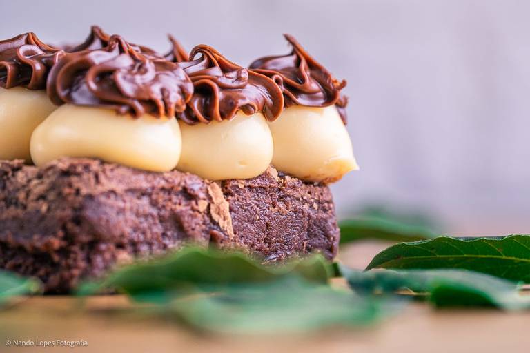 A sobremesa doce é um pedaço de bolo tipo brownie recheado com brigadeiro branco e confeitado de brigadeiro tradicional. Ao fundo textura de tijolos rústicos brancos, a base da mesa é de madeira e tem folhas verdes decorando a cena.