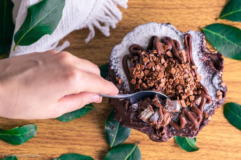 A sobremesa doce é uma Cesta de chocolate, com creme e confeitos de chocolate. Ao fundo textura de tijolos rústicos brancos, a base da mesa é de madeira e tem folhas verdes decorando a cena.