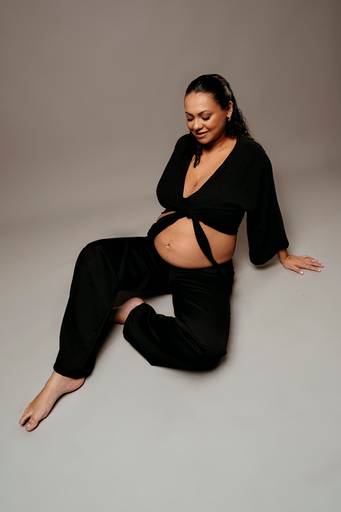 Ensaio gestante em estúdio, barriga a mostra, gestante esta sentada no chão com calça e top preto