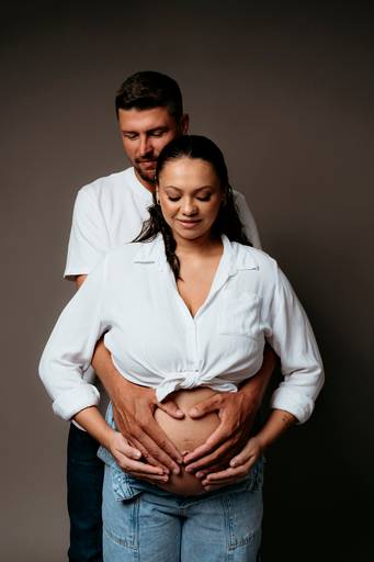Ensaio gestante em estúdio, gestante é abraçada  na barriga pelo marido e faz um coração com as mãos na barriga