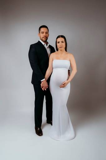 Ensaio gestante em estúdio, casal de mãos dadas, gestante está utilizando um vestido com técnica de  moulage branco e marido esta de terno