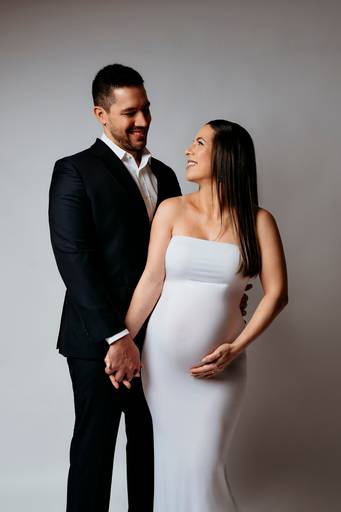 Ensaio gestante em estúdio, casal de mãos dadas olhando um para o outro, ela está com vestido feito com técnica de  moulage branco e marido esta de terno