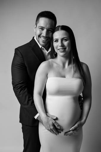 Ensaio gestante em estúdio, casal esta olhando para a foto, ele de terno atras da gestante e ela na frente usando um vestido branco feito com técnica de moulage