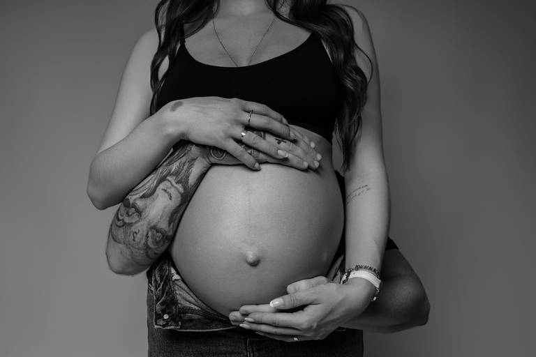 Ensaio gestante em estúdio, barriga a mostra braços tatuados do casal abraçam a barriga.