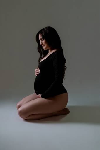 Ensaio gestante em estúdio, mulher de body preto de joelhos no chão olhando para a barriga