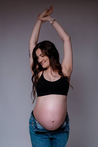 Ensaio gestante em estúdio, mulher sorri com os braços para cima barriga a mostra 