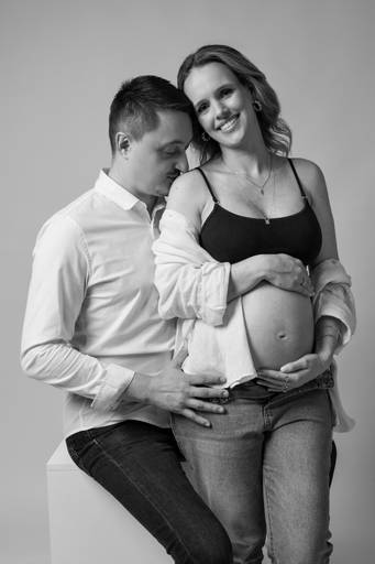 Ensaio gestante em estúdio, barriga a mostra, gestante abraça a barriga apoiada no marido, ele beija o ombro dela