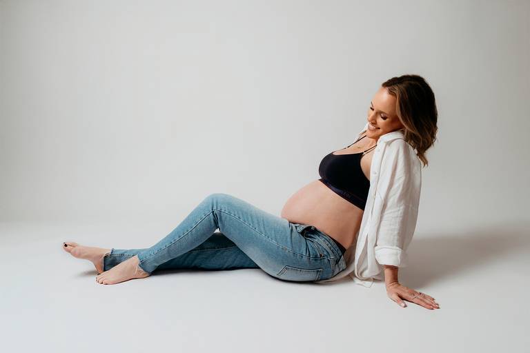 Ensaio gestante em estúdio, barriga a mostra, gestante sentada no chão olhando a barriga