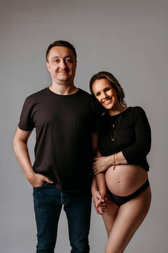 Ensaio gestante em estúdio, barriga a mostra, gestante abraça braço do marido, estão um do lado do outro de preto.