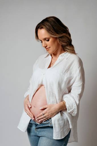 Ensaio gestante em estúdio, barriga a mostra, gestante abraça a barriga e olha para ela 