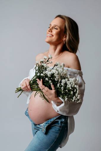 Ensaio gestante em estúdio, barriga a mostra, gestante abraça flores brancas que segura