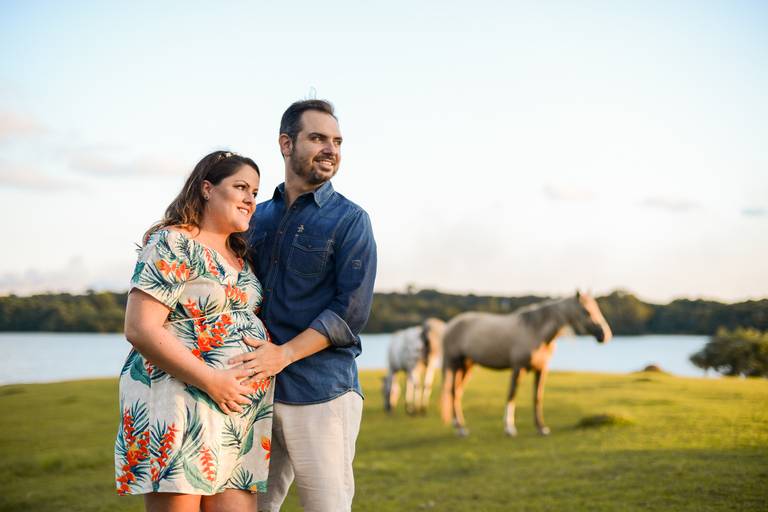 Ensaio de gestante fotografado pelo melhor fotógrafo de ensaios de campo largo e Curitiba. Casal olhando na mesma direção com as mãos na barriga e cavalos ao fundo