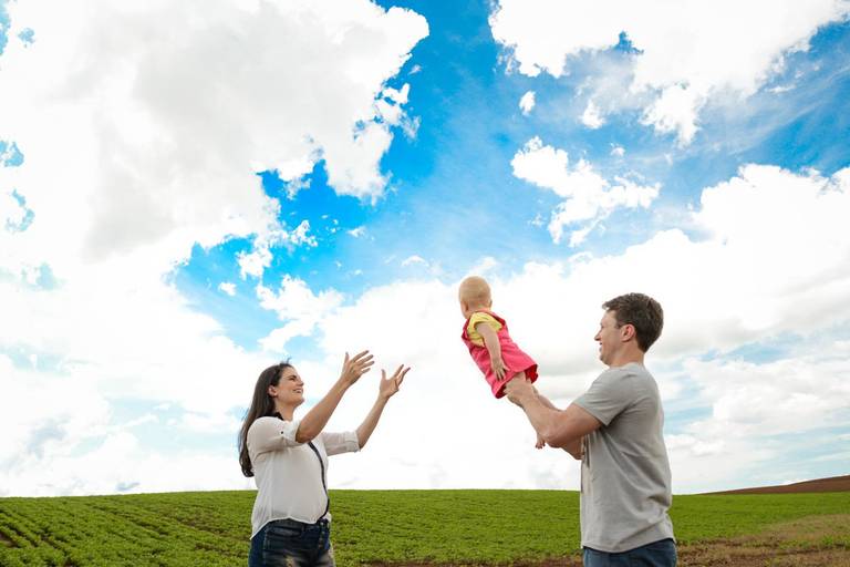 Família feliz, pai, mãe e filha fotografados pelo melhor fotógrafo de Curitiba e Campo Largo, Michel Druziki. Pai segurando a filha no campo com a mãe