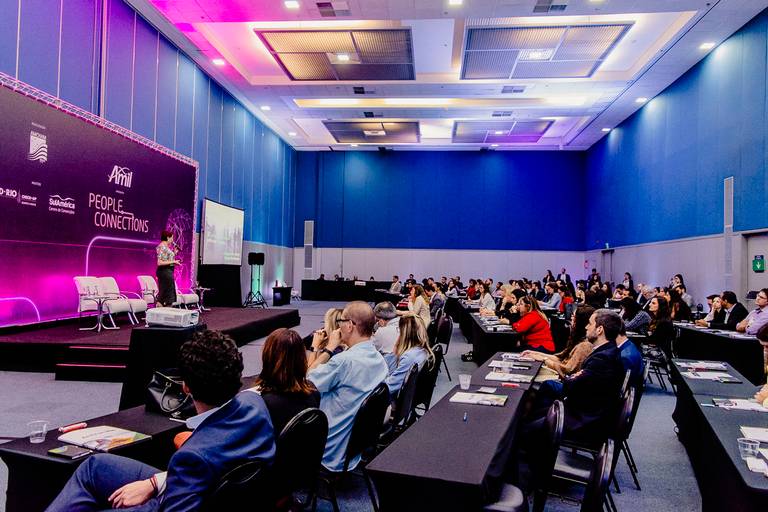 vista ampla de sala de palestras no evento feito em hotel em copacabana palco iluminado na cor lilás diante da plateia em filas de mesas e cadeira.