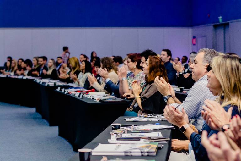 platéia organizada em mesas enfileiradas e batendo palmas para palestrante que faz sua apresentação.