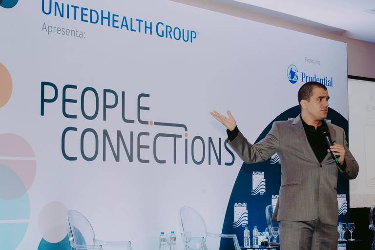 evento corporativo produzido pela AMCHAM em hotel em copacabana. Palestrante vestido de blazer cinza e camisa preta segurando microfone e apontando para a tela onde apresenta o conteúdo de sua palestra