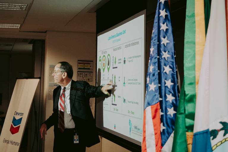 evento corporativo na empresa chevron no centro do rio de janeiro. Executivo da empresa usando terno preto e gravata vermelha e branca mostra na tela resultados e tabelas de desempenho da empresa. bandeiras dos EUA e do Brasil estão ao lado da tela