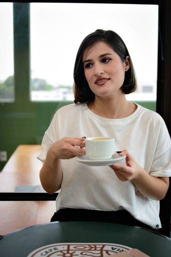 ensaio externo Niterói de empreendedora social media e designer. A empreendeora segura uma xícara de café e olha para o lado direito com leve sorriso. A designer veste blusa branca básica e está em uma mesa em um bistrô em Nitéroi.