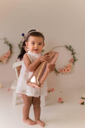 Fotografia de Smash the cake para comemorar primeiro ano do Bebê. Bebê segurando o número 1, vestindo vestido branco de renda com flores ao fundo