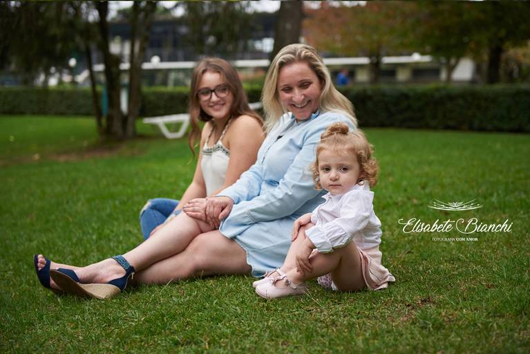 Criança, irmã e mãe sentadas no gramado, em ensaio infantil externo, em Caxias do Sul.