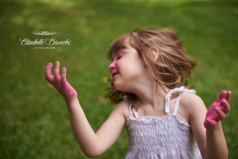 Criança em movimento, tirando os cabelos do rosto, com as mãos sujas de pó cor de rosa, em ensaio infantil externo, em Caxias do Sul.