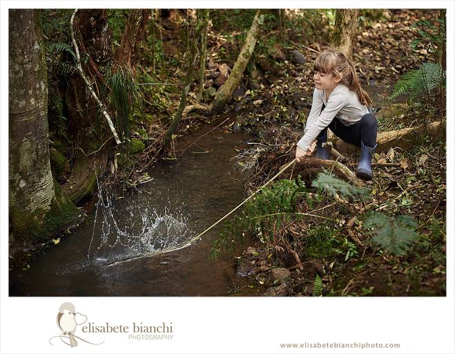 Criança brincando em córrego, com uma vareta, batendo na água, em ensaio infantil externo, entre Caxias do Sul e Farroupilha. Essa foto recebeu prêmio  de fotografia no Amarelo Awards.